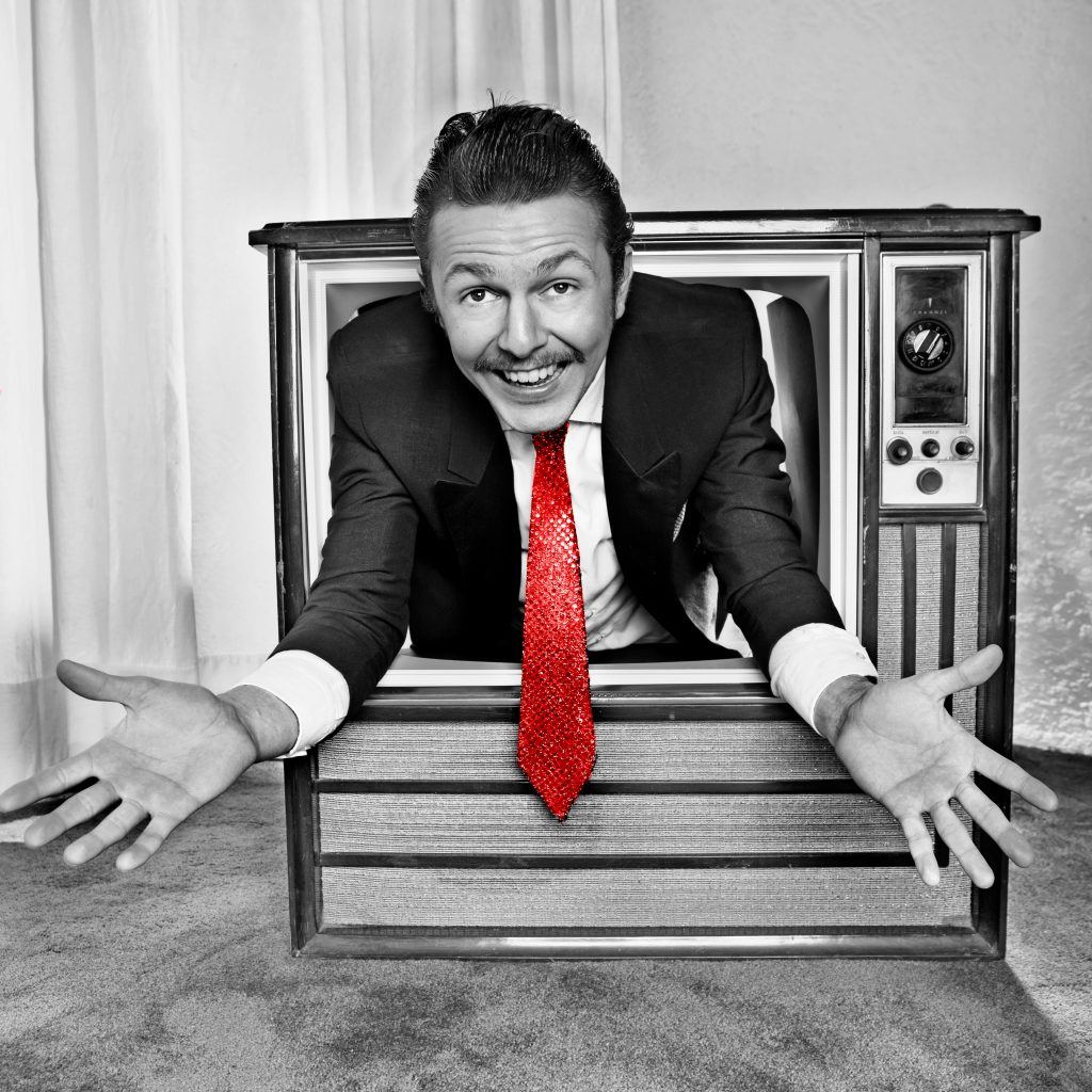 Homme en costume et cravate rouge émergeant d'un poste de télévision vintage, bras ouverts, illustration de la réactivité de l'agence DUNK!